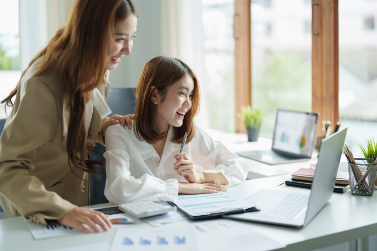 Two woman sit at desk feel euphoric win online lottery, happy asian woman overjoyed get mail at laptop being promoted at work, biracial girl amazed read good news at computer.